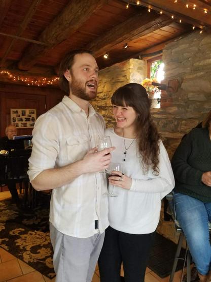 A man with short brown hair and a beard, wearing a light-colored button-down shirt and grey pants, stands next to a smiling woman with long brown hair wearing a white long-sleeved shirt and black pants, both holding wine glasses in a rustic room with exposed wooden beams.