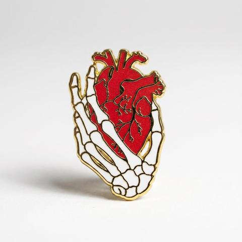 An enamel pin featuring a skeletal hand holding a red anatomical human heart with gold outline details, set against a plain light gray background.