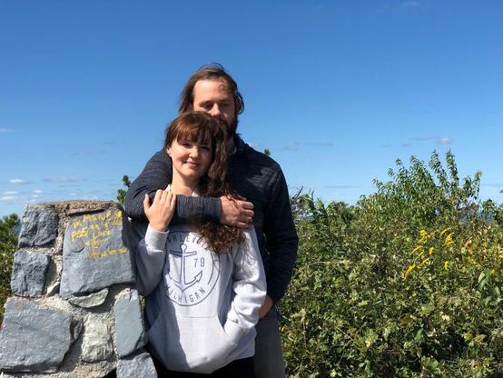 A man embraces a smiling woman from behind as they pose outdoors against a clear blue sky next to a stone marker.