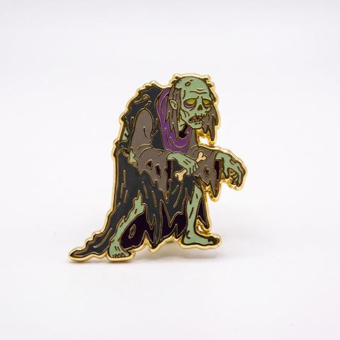 An enamel pin depicting a hunched, decaying green zombie in tattered dark robes, holding a bone.