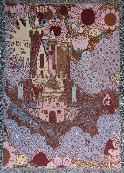 A woven tapestry blanket featuring a whimsical storybook castle surrounded by large flowers, starry clouds, butterflies, mushrooms, and a stylized sun with a face.